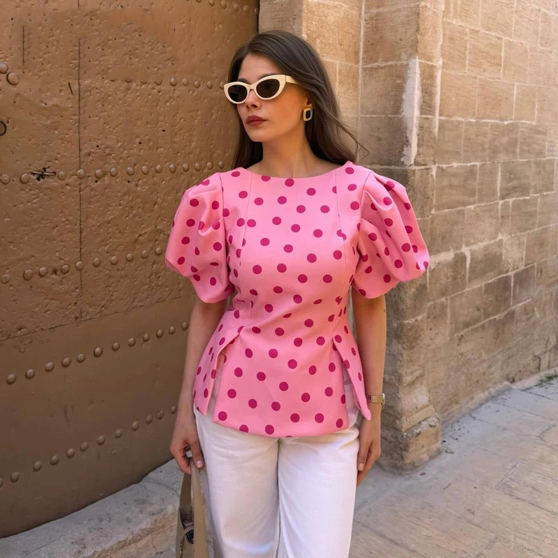 Pink Polka Dot Puff Sleeve Blouse 1 Woman wearing a vibrant pink Polka Dot Puff Sleeve Blouse with large puff sleeves and a side-slit peplum hem, styled with white high-waisted trousers and cat-eye sunglasses.